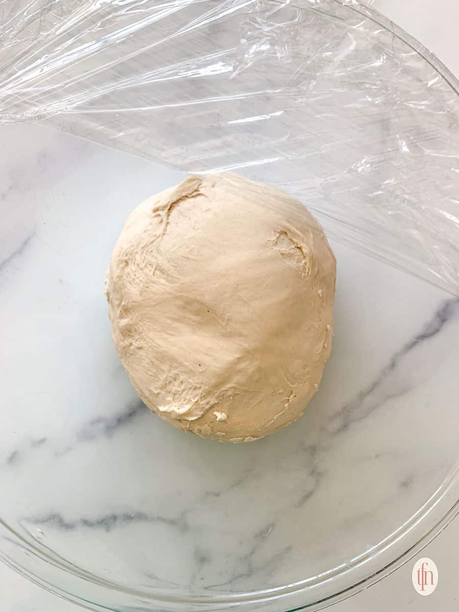 a roll of dough in a glass bowl being covered with plastic wrap to rest.