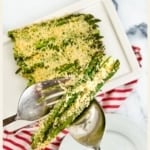 A vertical image with the title "ASPARAGUS casserole recipe" at the top. The image shows a silver fork and spoon holding a serving of baked asparagus covered in a cheesy topping. A white platter with the remaining casserole is blurred in the background. The website "TheFeatheredNester.com" is at the bottom.