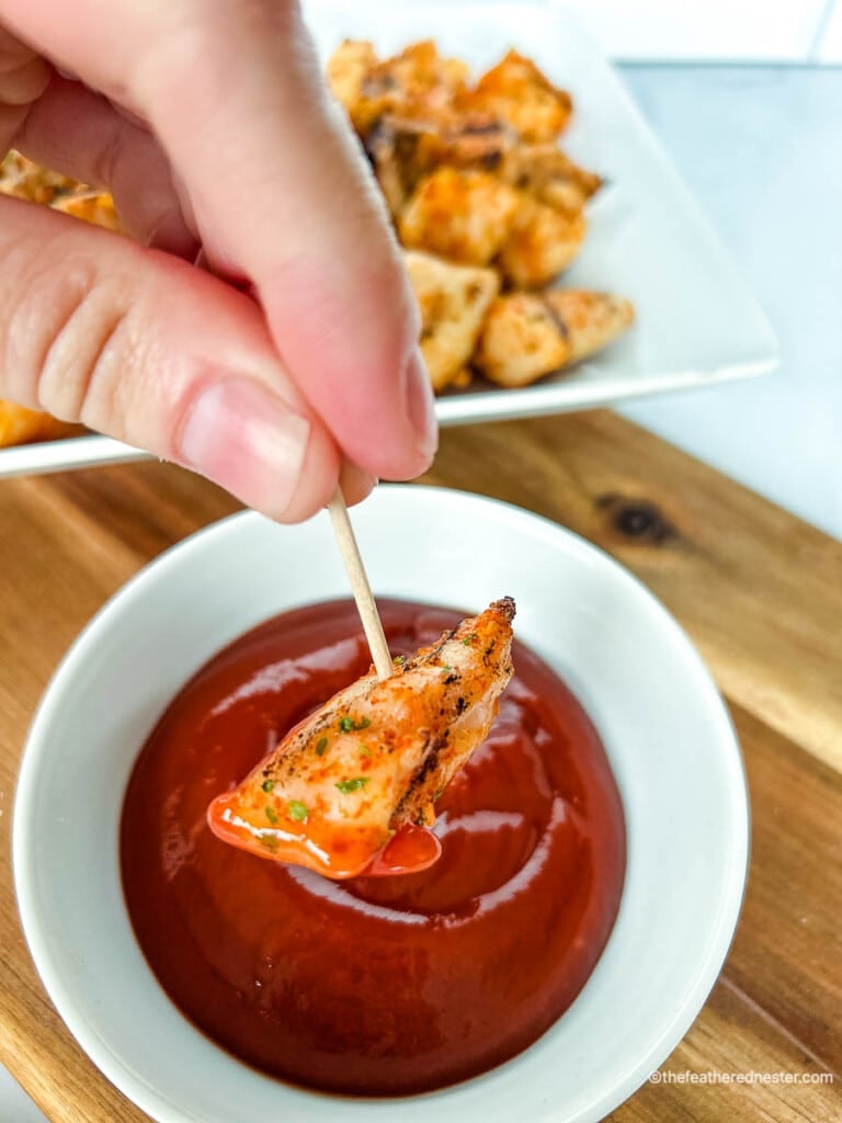 Small piece of cooked meat on a toothpick being dipped into red sauce.