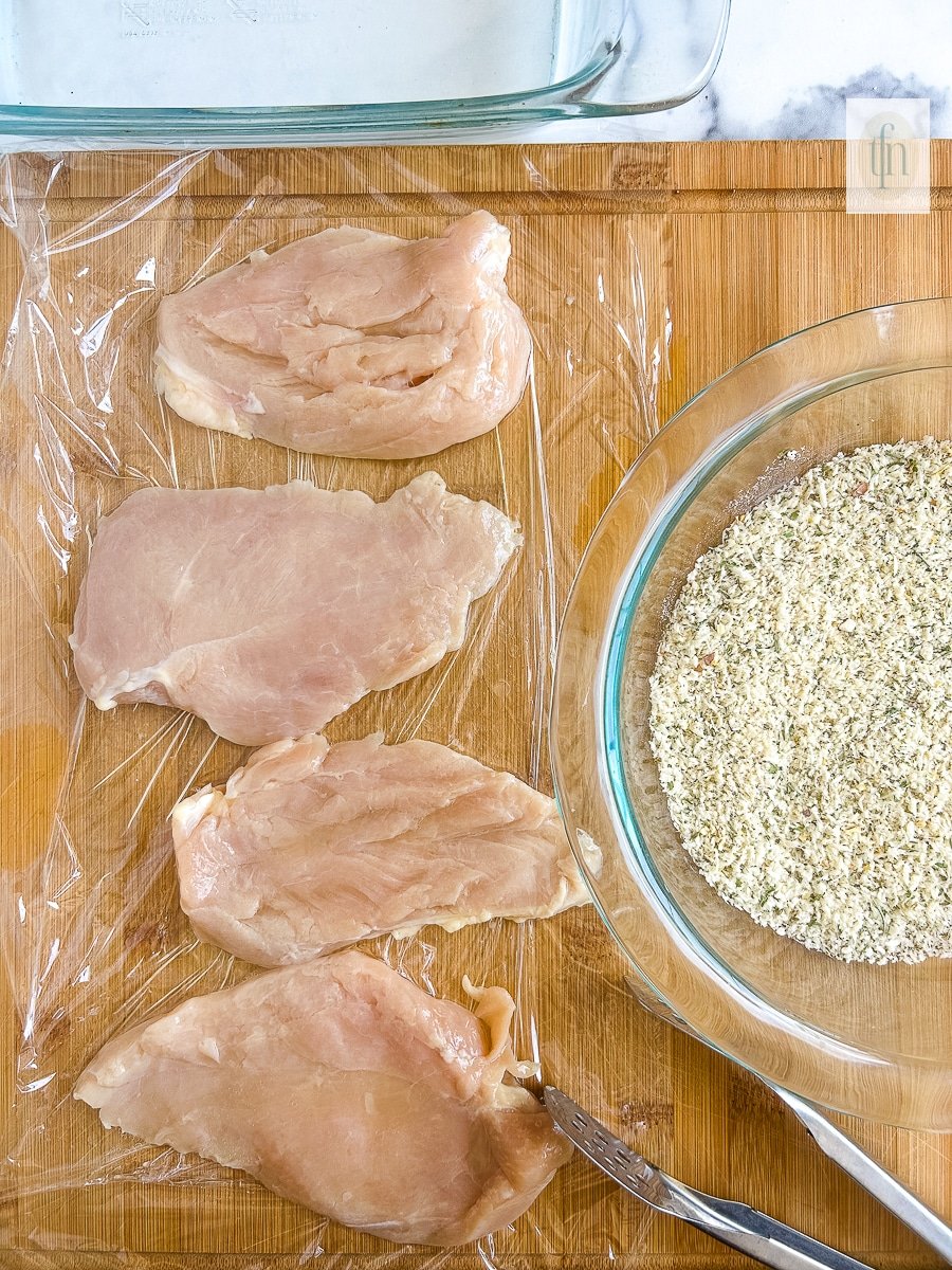 Several pieces of uncooked boneless skinless chicken breast on a piece of plastic wrap, next to a bowl of bread crumbs.
