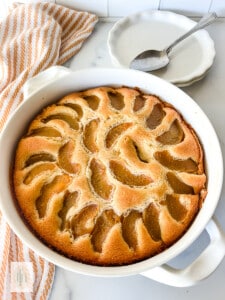 Peach cobbler pound cake baked in a round white baking dish. Stack of dessert plates and spoons sitting behind the pound cake.