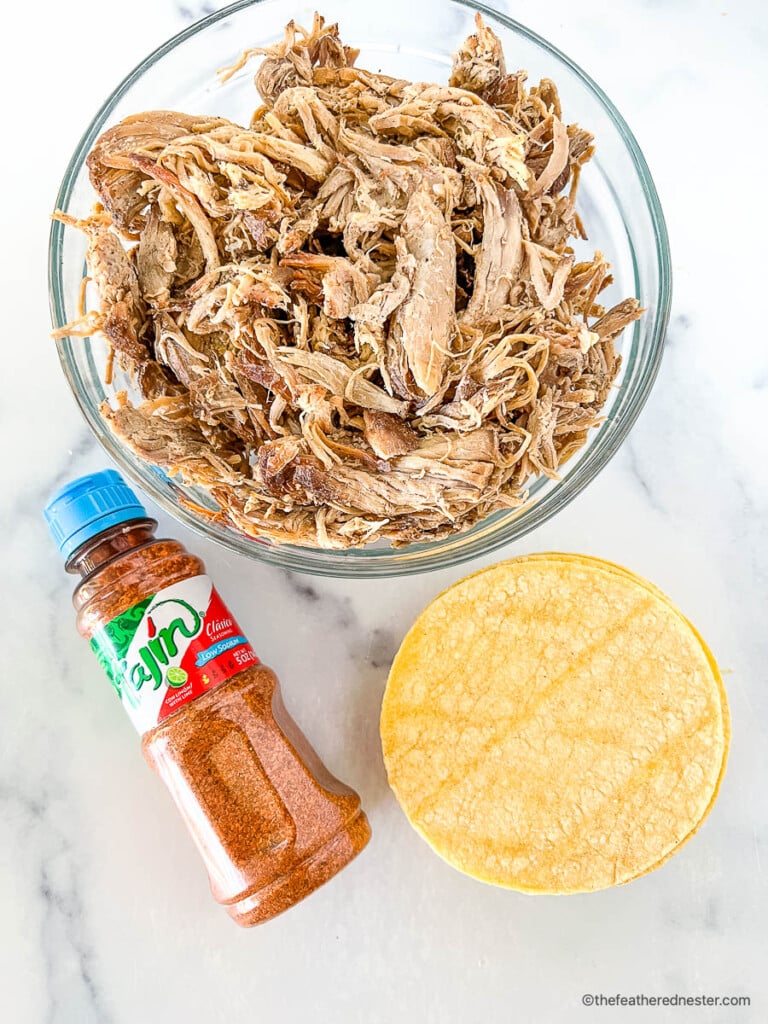 Bowl of leftover Mexican pulled pork next to a bottle of Tajin chile lime seasoning and street taco tortillas.