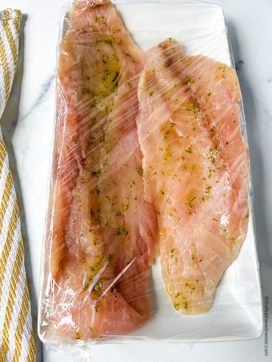 Two large Hawaiian rock cod filets on a white plate tightly wrapped in plastic wrap.