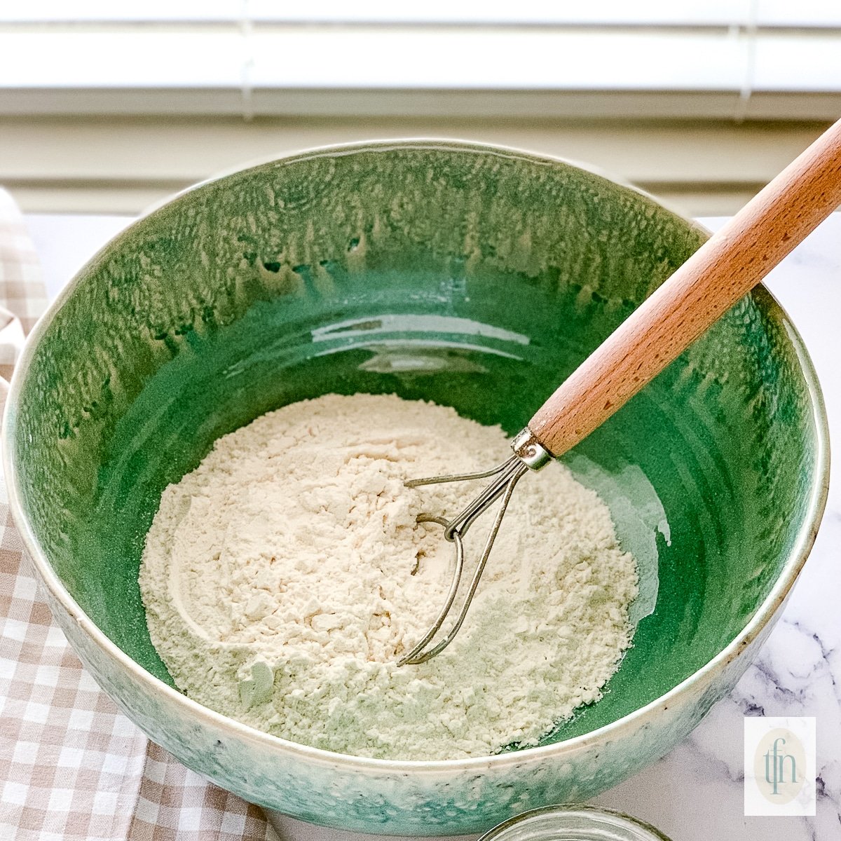 A green bowl with dry ingredients and a dough whisk to stir them.