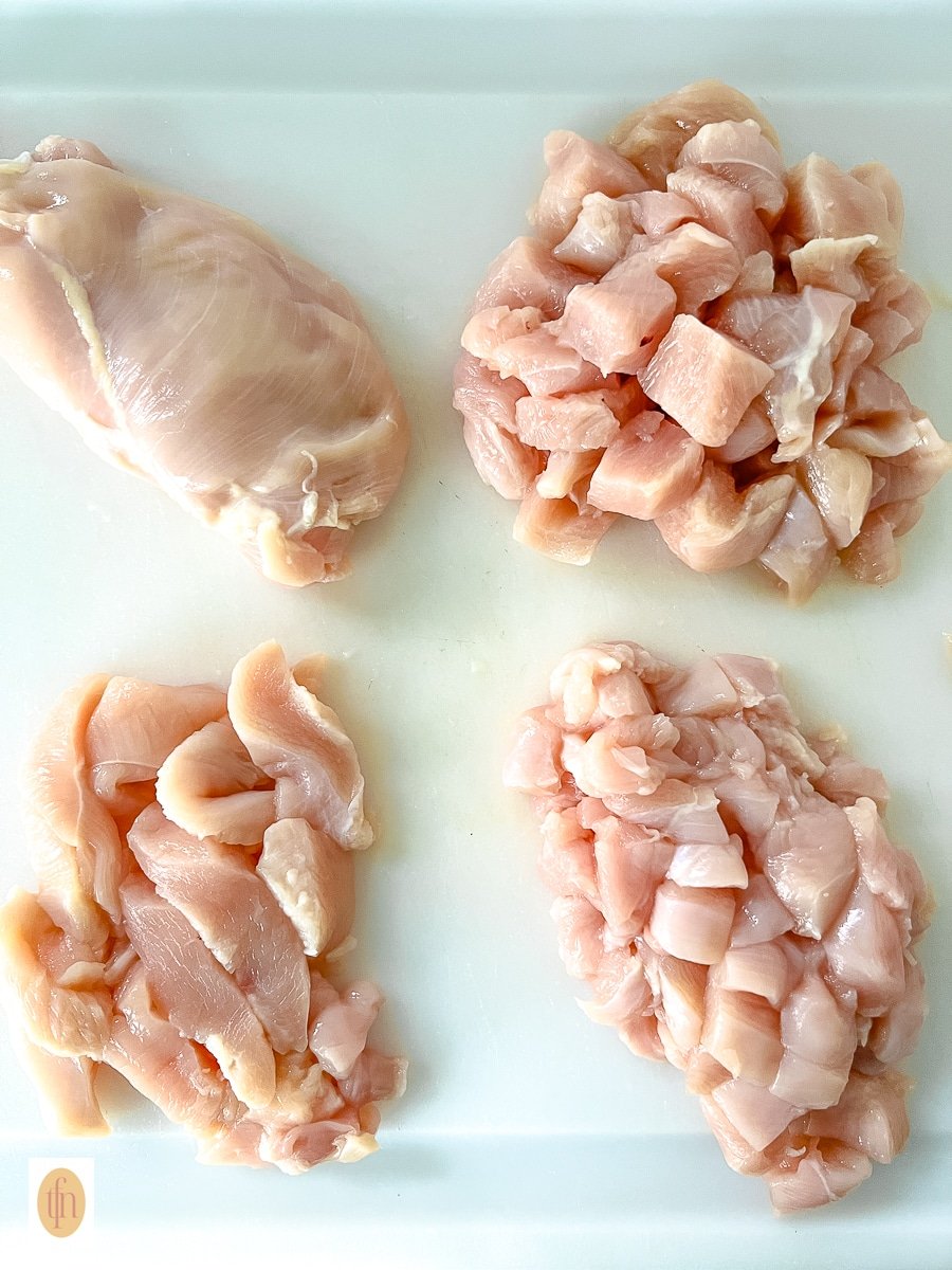 Four raw chicken breasts on a cutting board. One is a whole breast, one is chicken breast cubes, one is chicken strips, and one is diced chicken breast.