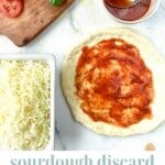 Sourdough discard pizza crust topped with pizza sauce sits next to a dish full of shredded white cheese.