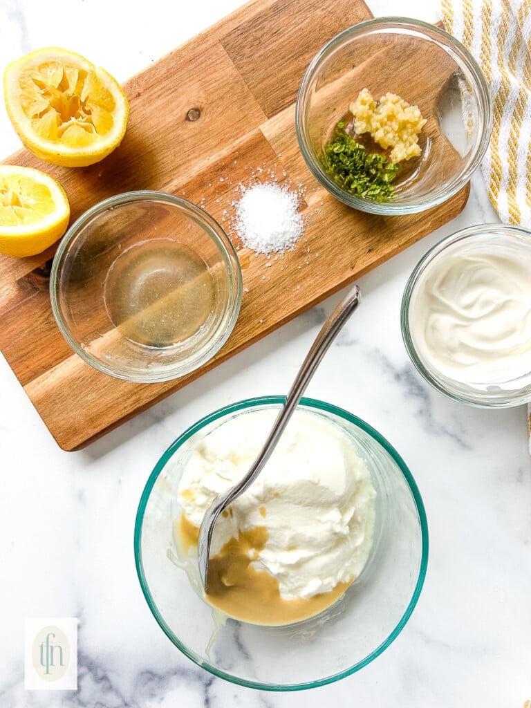 Stirring tahini into a bowl of Greek yogurt. Small bowls with minced garlic and lemon juice sit behind the bowl.