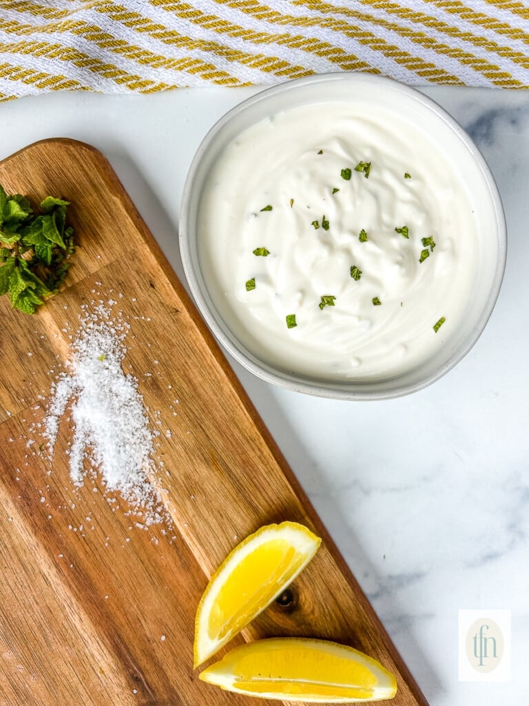 Small dish of shawarma sauce next to a cutting board with lemon wedges, salt, and chopped cilantro.