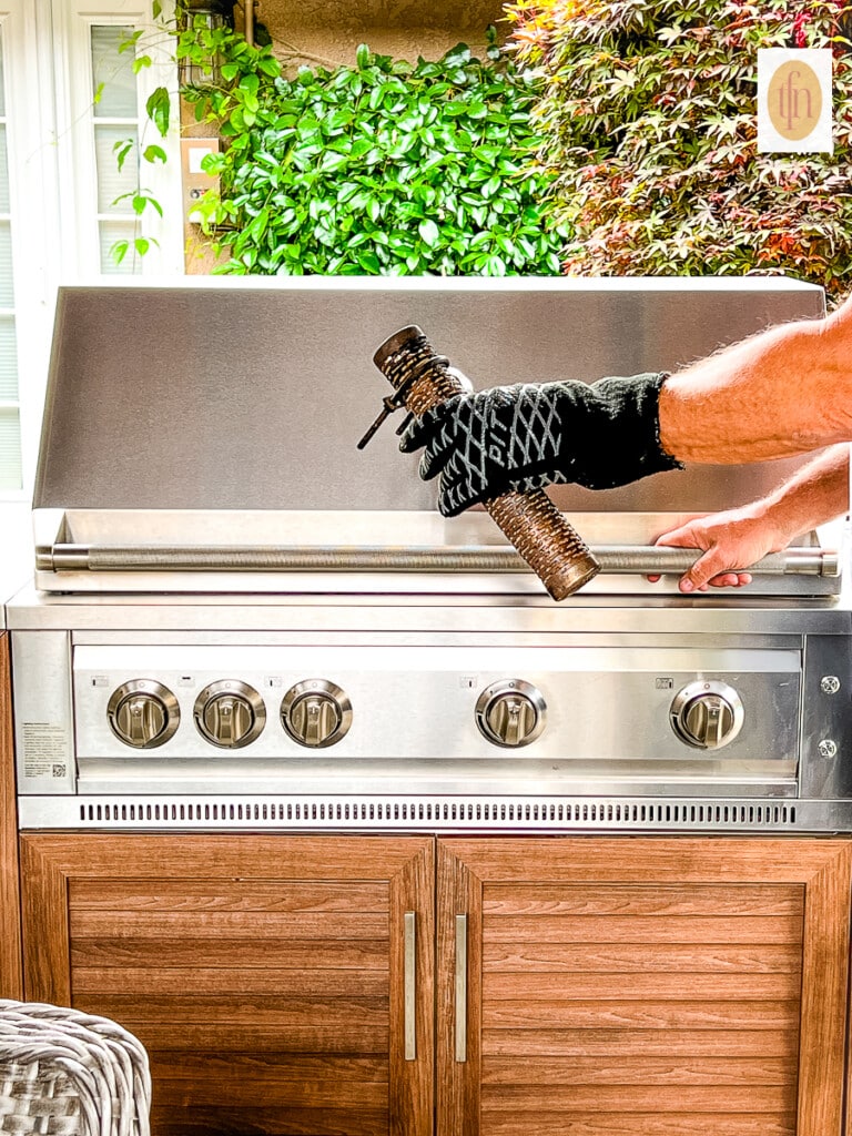 Man's gloved hand holding a pellet smoker tube next to a gas grill.