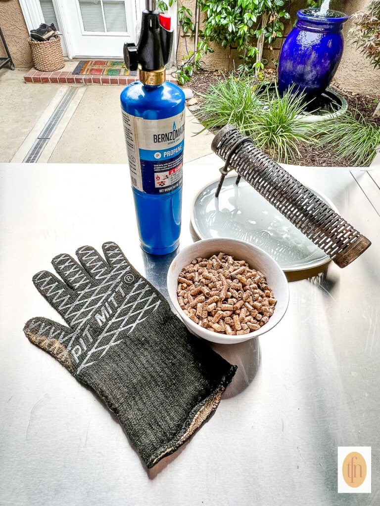 Grilling glove on a table with grill pellets, a blue propane torch and smoke tube.