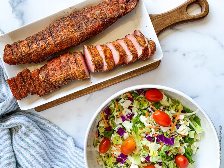 A wooden board with sliced, seasoned pork tenderloin and a bowl of colorful salad featuring tomatoes, carrots, and cabbage on a marble surface. A striped cloth is placed next to the dish.