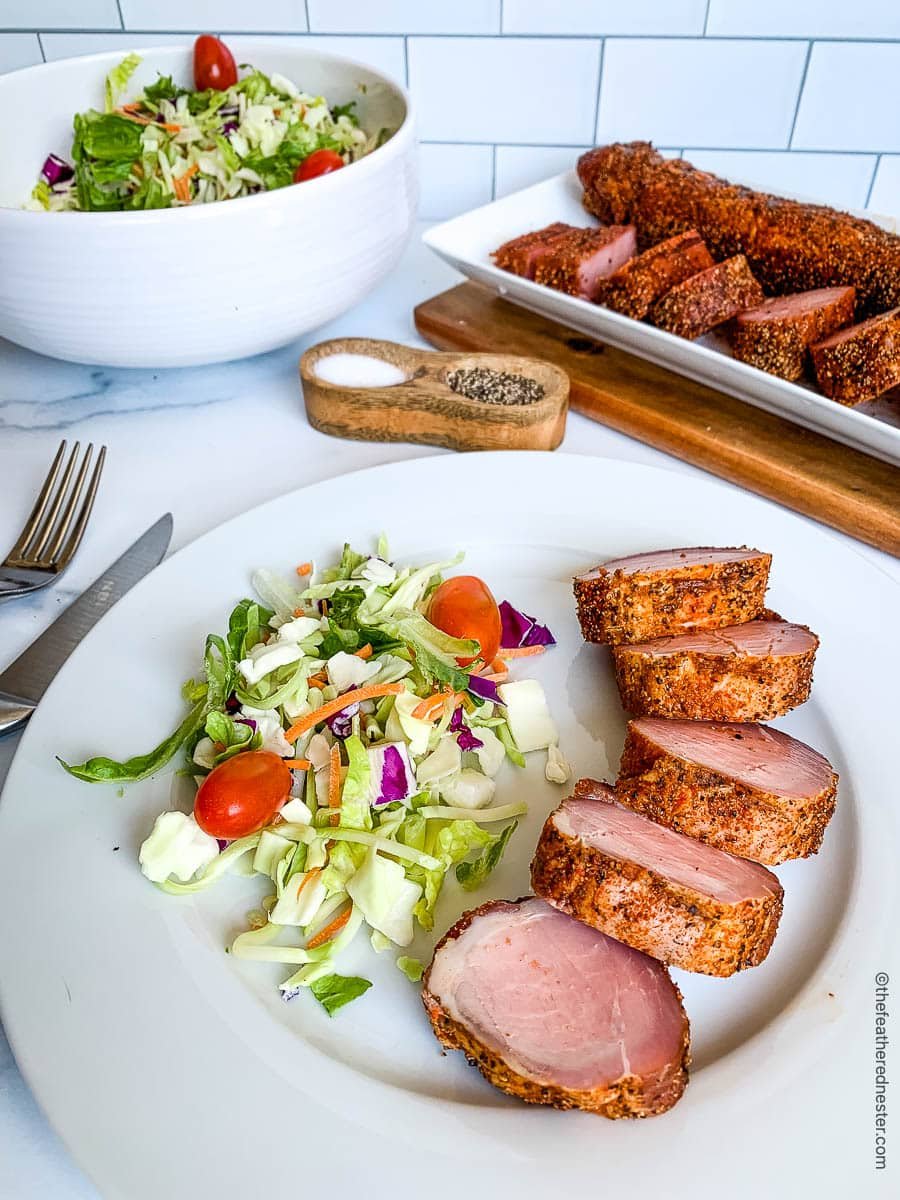 Slices of smoked pork tenderloin with salad on a dinner plate and on a platter in the background.