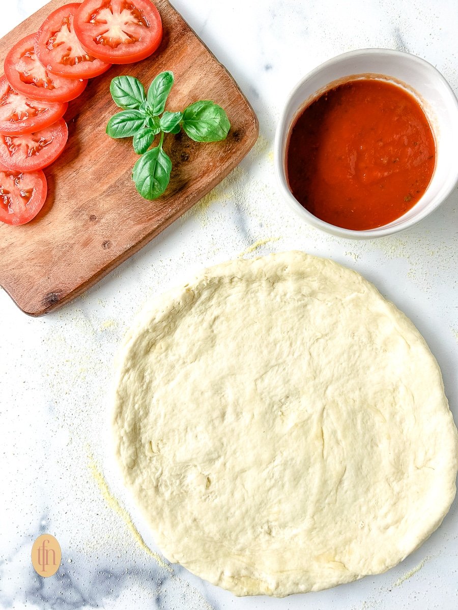 Sourdough discard pizza dough on a floured counter top next to a bowl with pizza sauce, and tomato slices, and fresh basil leaves on a cutting board.