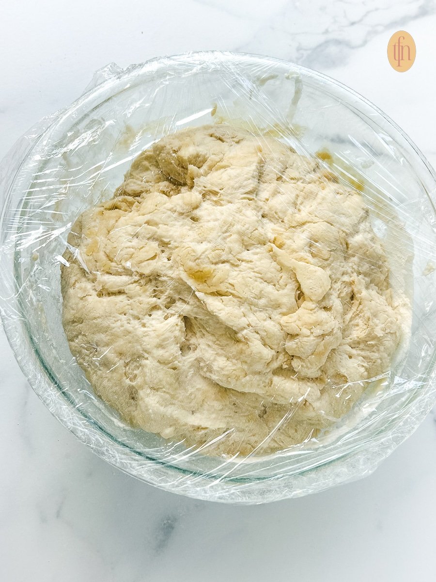 Bowl of Sourdough Discard Pizza Dough rising in a glass bowl covered with plastic wrap.