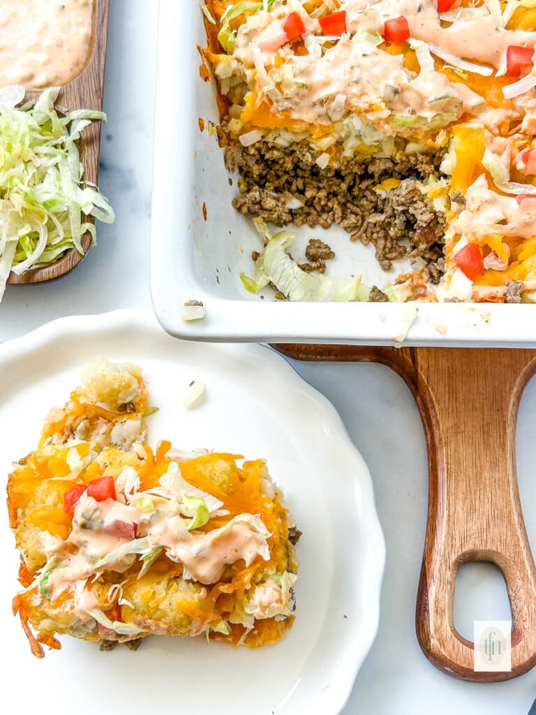 Plated serving of cheeseburger tater tot casserole next to the remaining casserole in a baking dish.