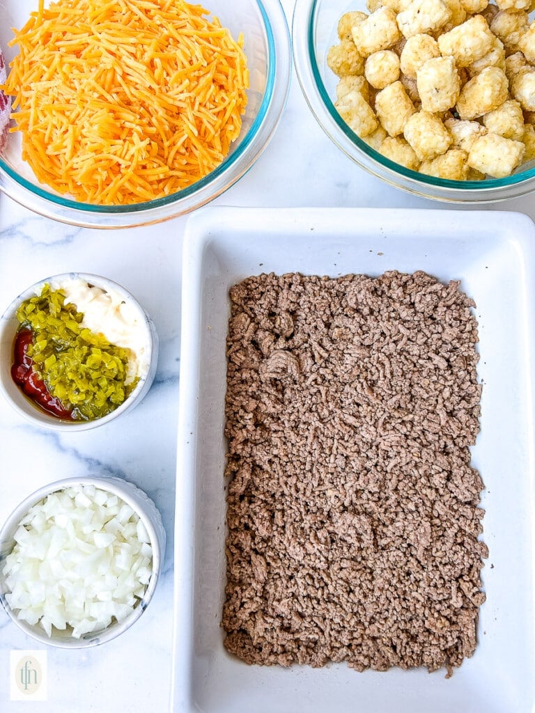 Cooked ground beef in a white baking dish surrounded by smaller bowls of diced onion, bell pepper, shredded cheese, and tater tots.