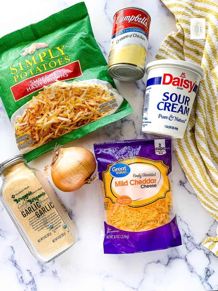 Packages of shredded potatoes, condensed soup, sour cream, and cheddar cheese on a counter next to garlic powder and onion.