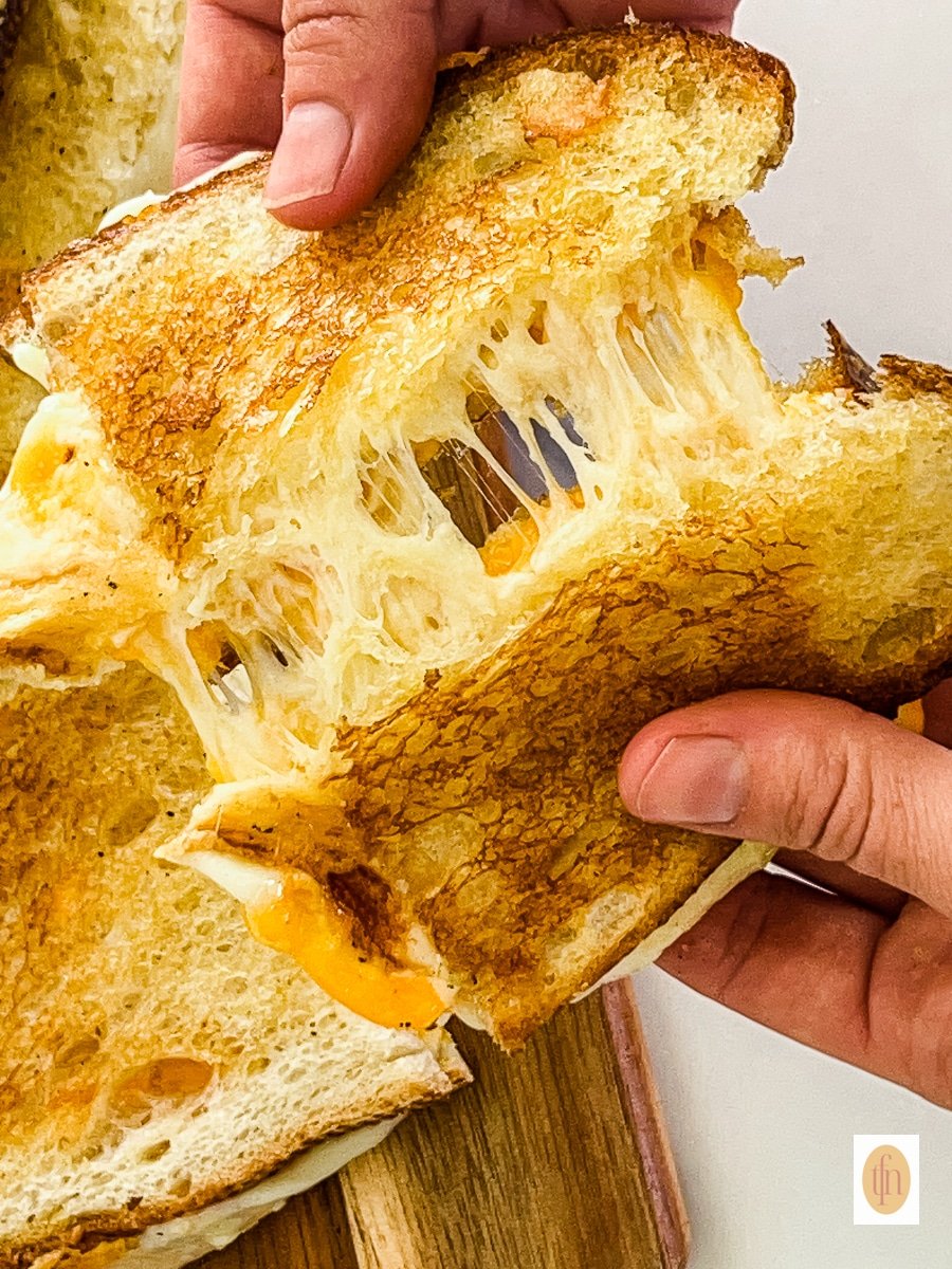 Man's hands pulling apart two halves of a melted cheese sourdough sandwich.