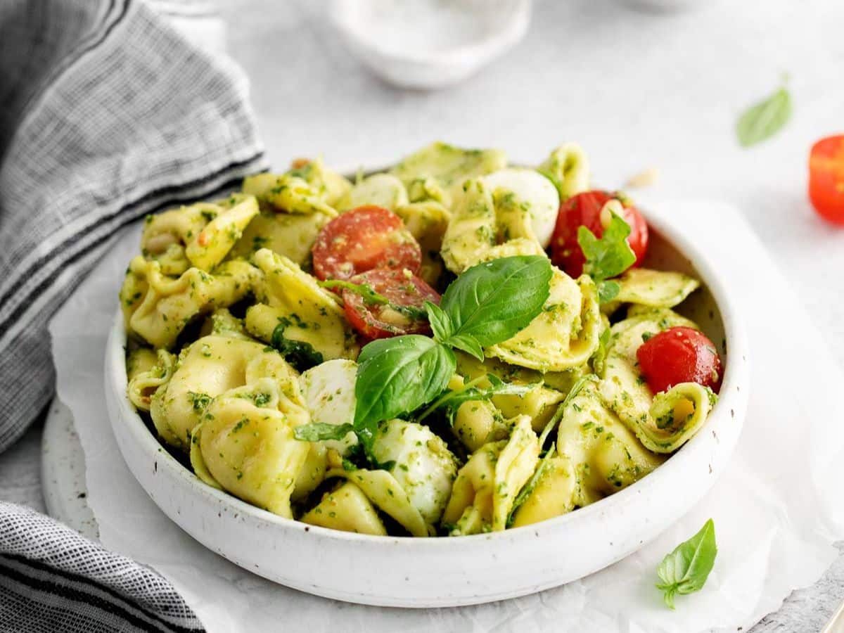 Tortellini, cherry tomatoes, and mozzarella balls, all covered with pesto sauce in a white pasta bowl.