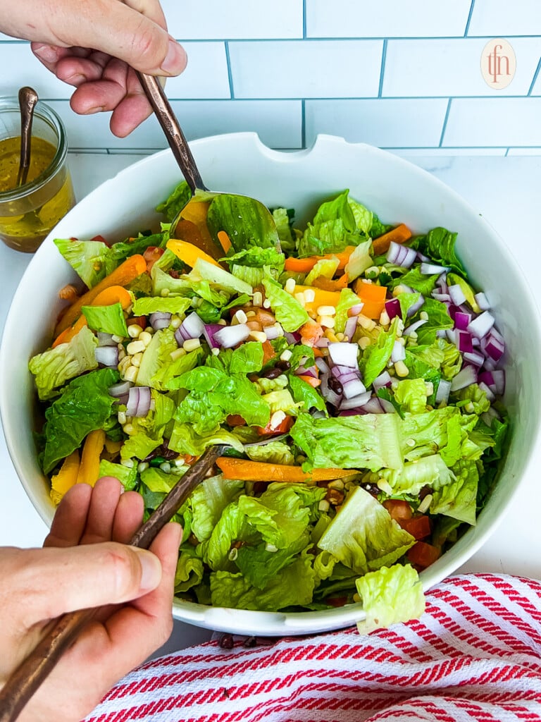 Tossing romaine lettuce, chopped vegetables, and dressing in a large serving bowl.