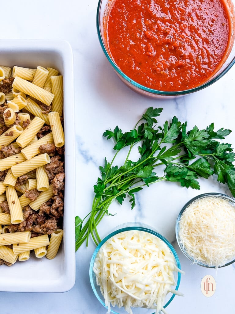 Italian sausage and rigatoni pasta in a white baking dish next to bowls with marinara, parmesan, and mozzarella.