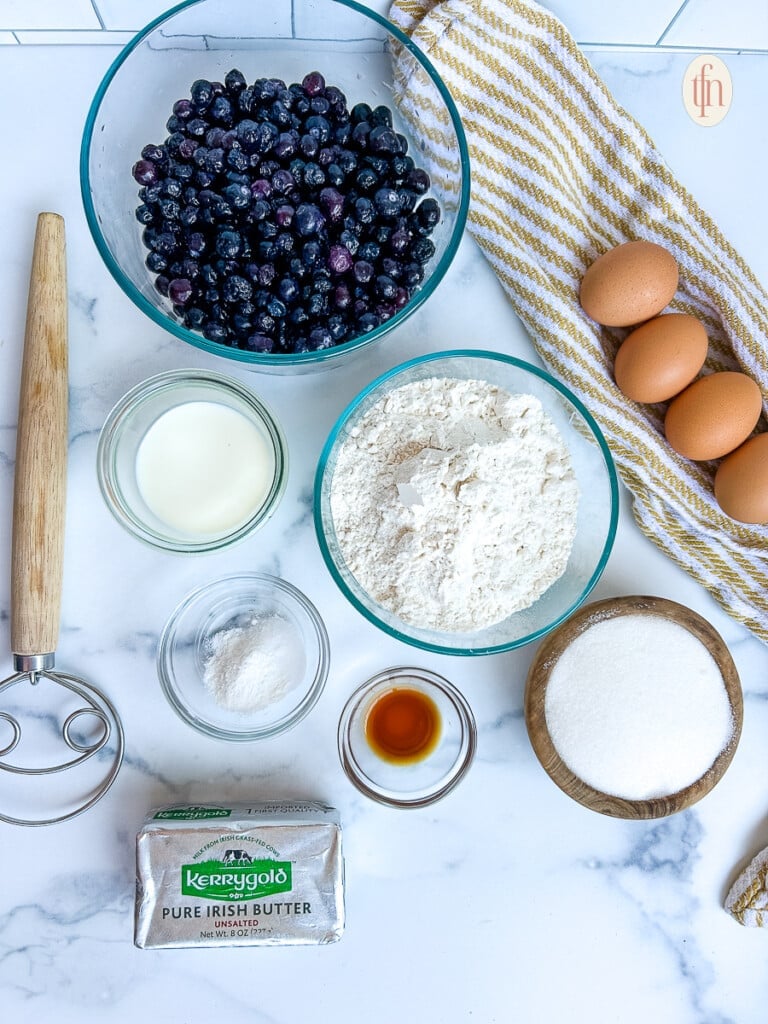 Glass bowls with sugar, all-purpose flour, milk, and vanilla extract sitting next to other blueberry cake ingredients.
