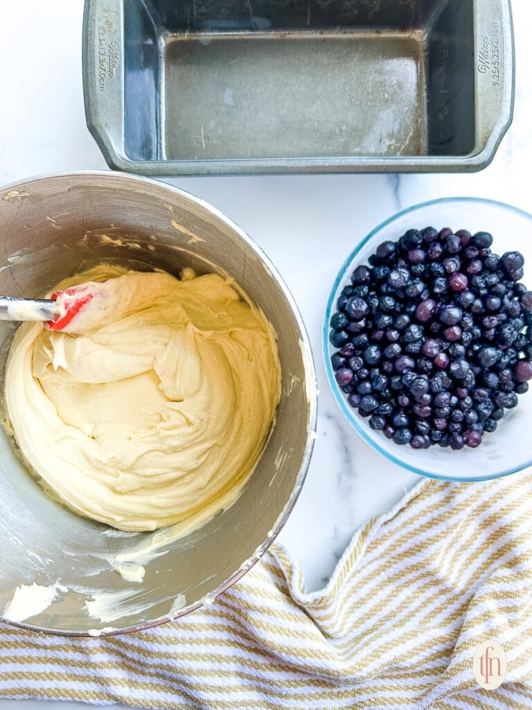 overhead shot of an empty loaf pan, bowl of frozen blueberries, and a stand mixer bowl with pale yellow batter in it.