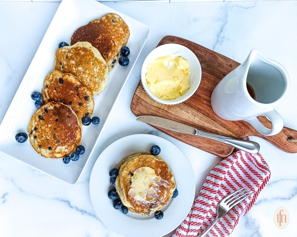 Serving platter and individual plated serving of muffin mix pancakes next to dishes with butter and maple syrup.