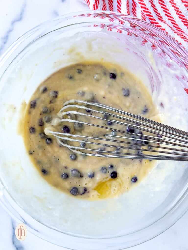 Whisking blueberry pancake batter in a glass mixing bowl.