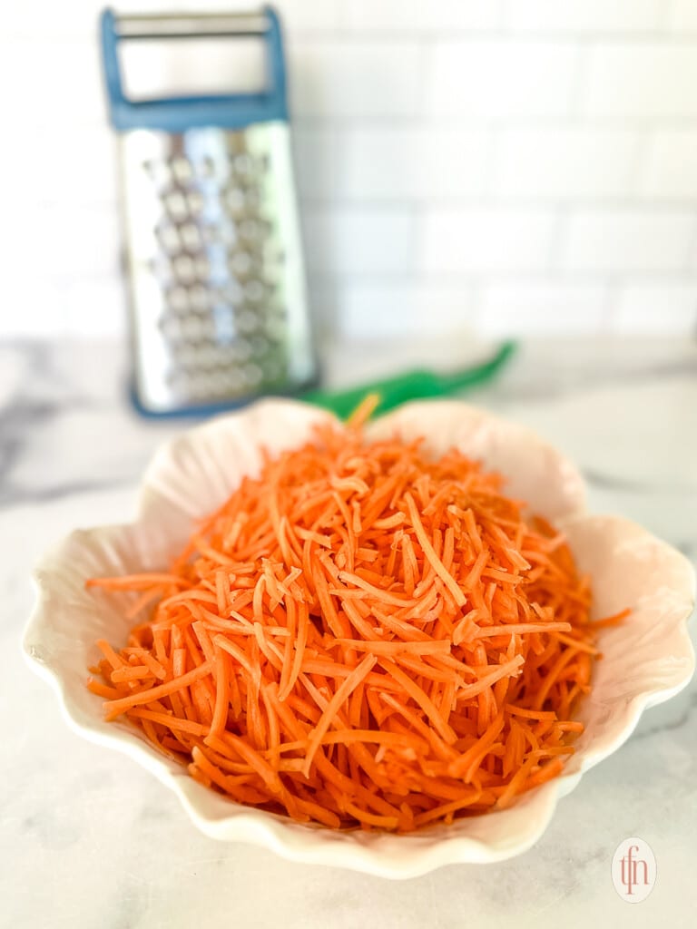 Grated carrots in a small white bowl, and a box grater in the background.