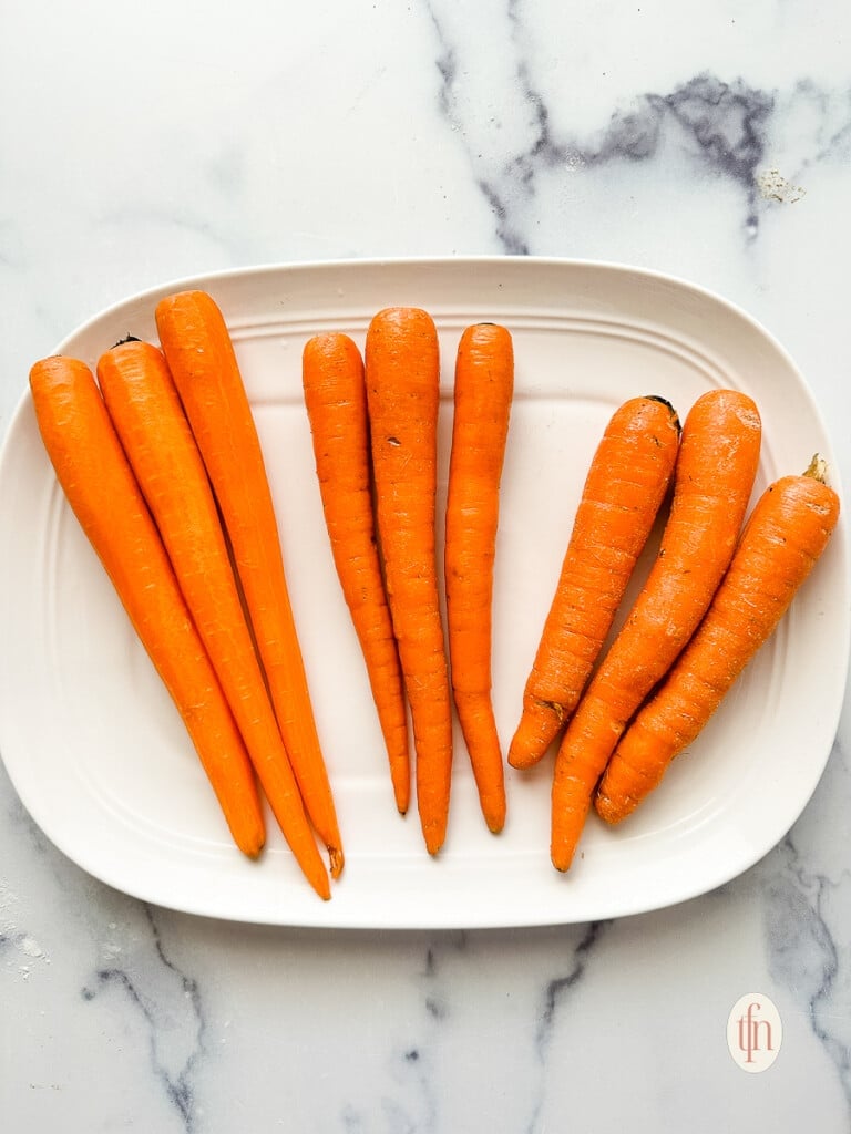 Nine long, thin, orange root vegetables on a white serving plate. Three of them have been peeled.