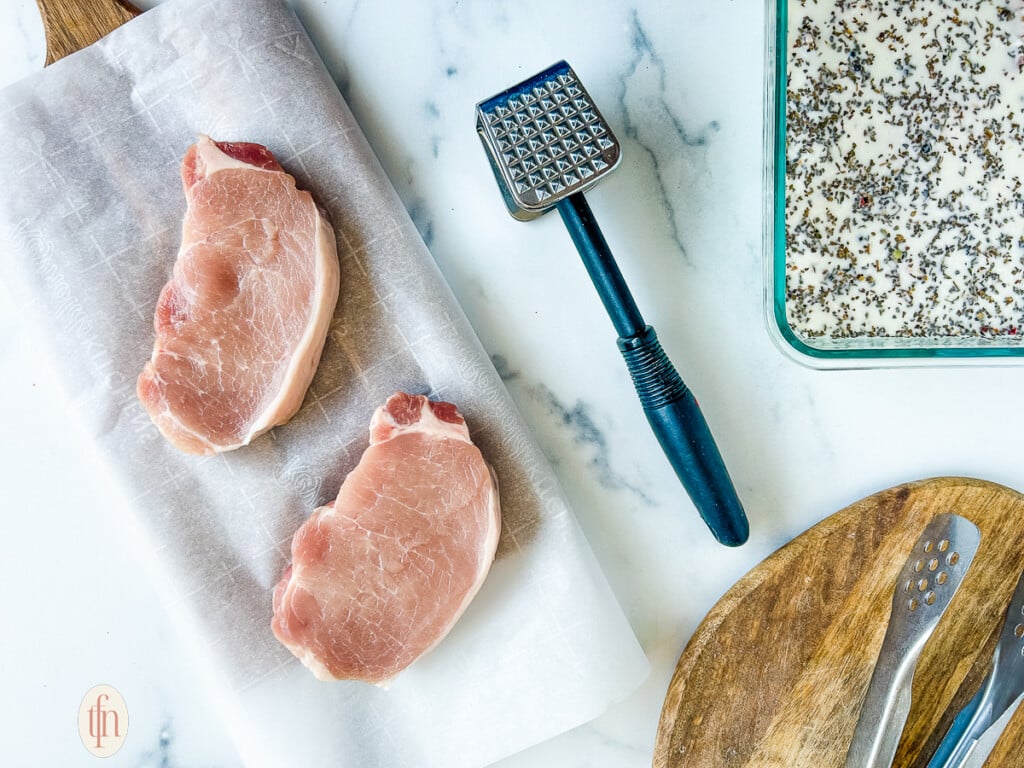 Two raw pork chops on a cutting board next to a meat mallet.