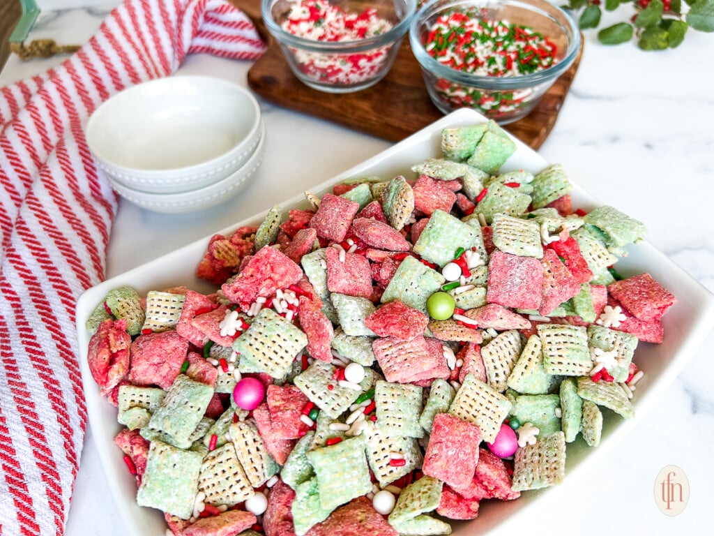 Large white dish containing Christmas muddy buddies next to smaller bowls filled with sprinkles on a white marble countertop.