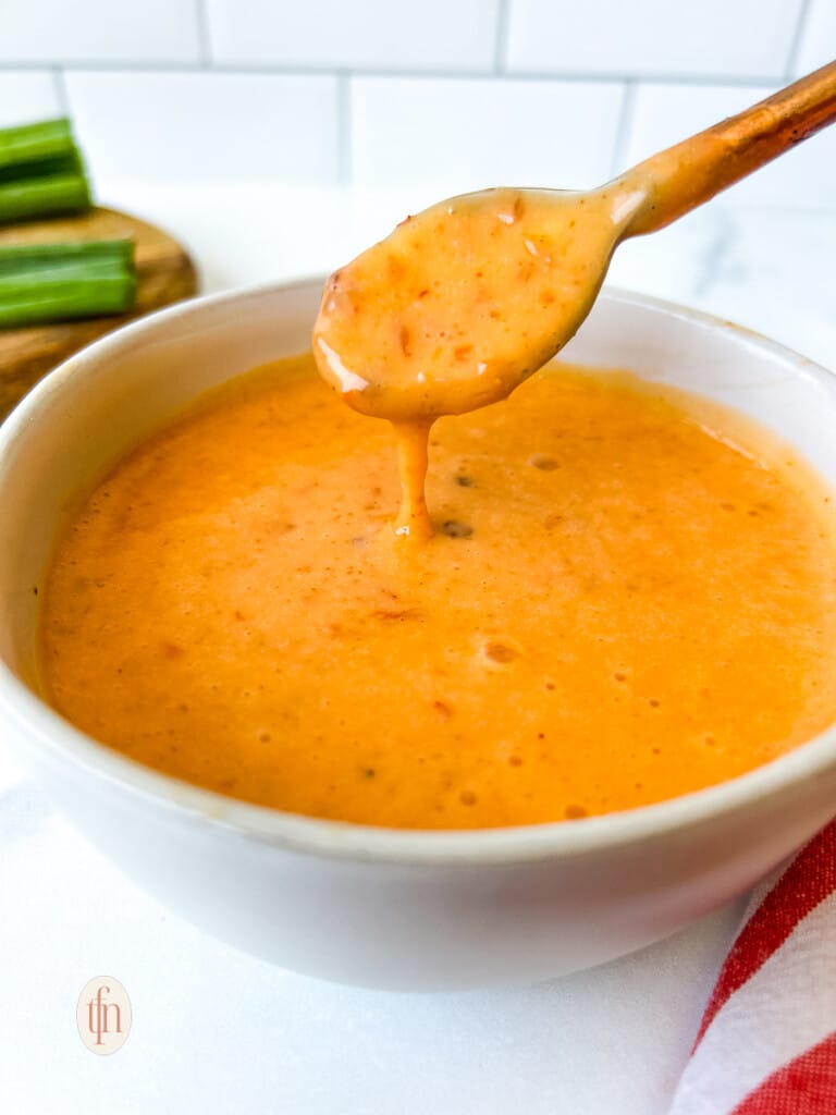Thick and creamy bang bang sauce dripping from a small spoon into a white bowl with more of the spicy dipping sauce.