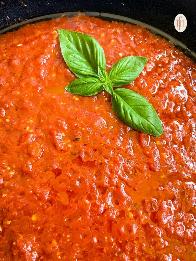 Tomato sauce in a cast iron pan on with a sprig of basil on top on a white background.