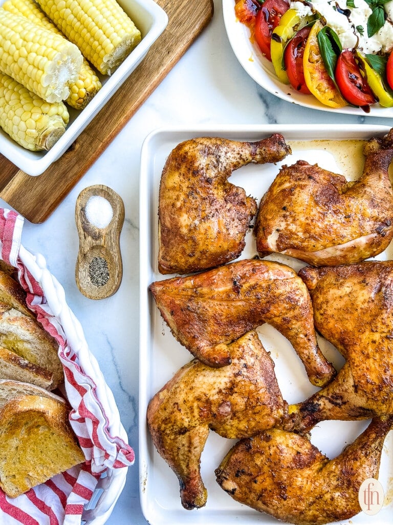 Pretty arranged image of chicken on a white platter, corn on the cob, bread slices, and tomatoes.