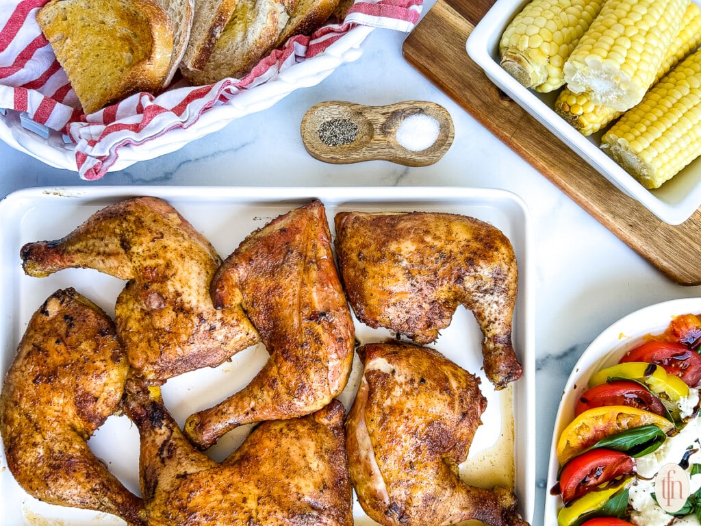 Pretty arranged image of chicken on a white platter, corn on the cob, bread slices, and tomatoes.
