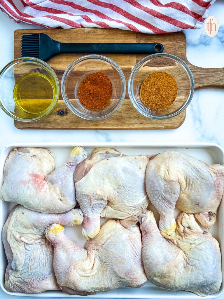Ingredients for smoked chicken quarters including raw chicken on a large platter and spices in small bowls on a wooden cutting board.