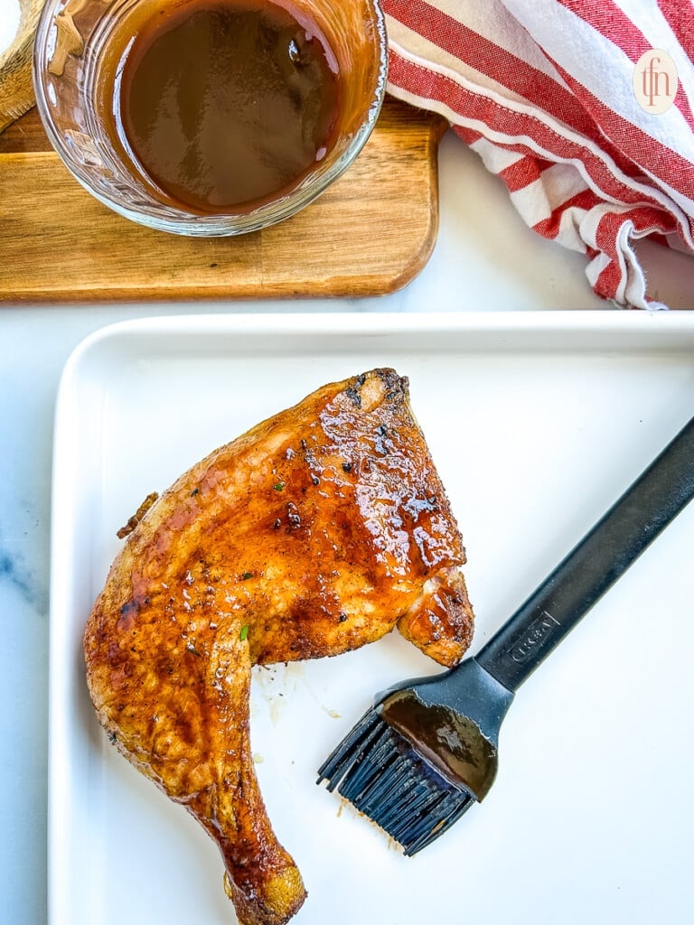 One chicken quarter on a white plate with a black silicone brush coated in barbecue sauce sitting next to it.