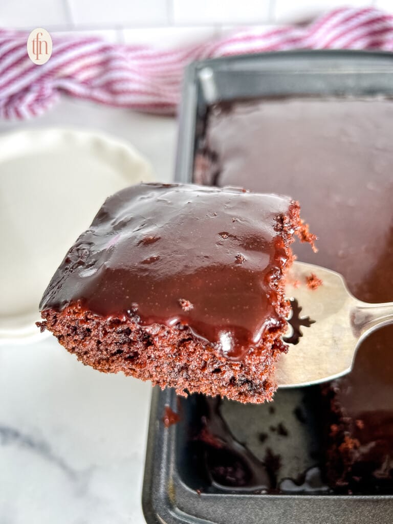 Silver serving spoon holding slice of cake over the pan of chocolate cake.