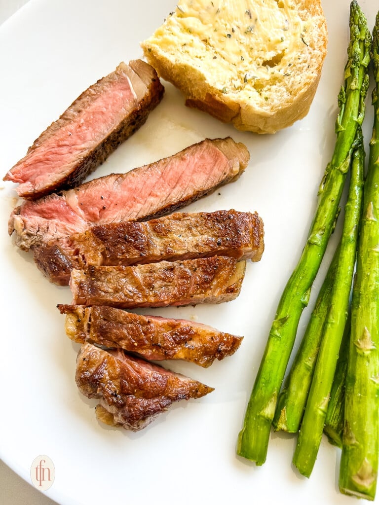 Slices of grilled NY strip steak on a dinner plate with asparagus spears and a slice of buttered bread.
