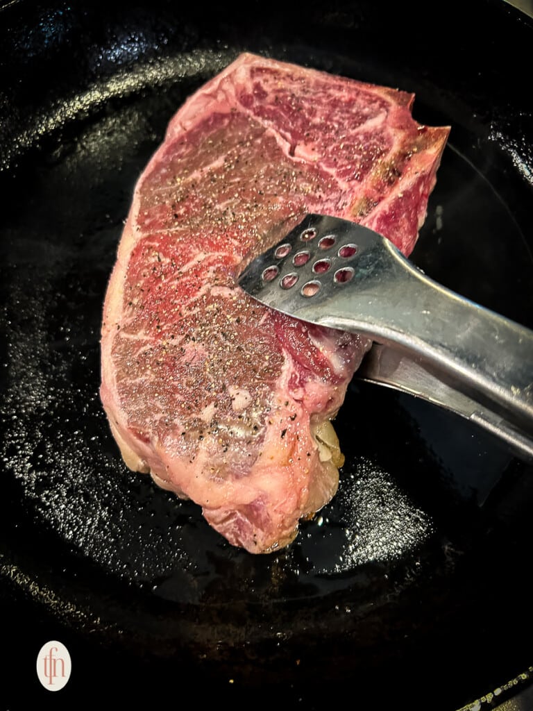 Using tongs to turn over a bone-in NY strip steak as it cooks in a skillet