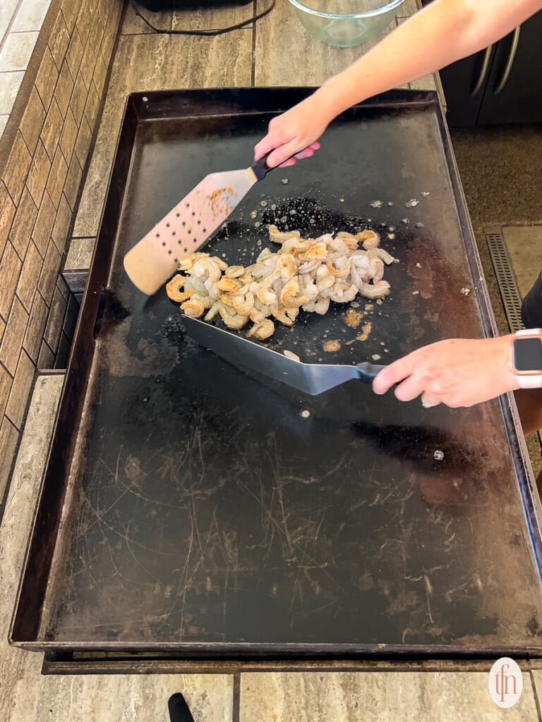 Flipping raw shrimp with metal spatulas on a griddle.