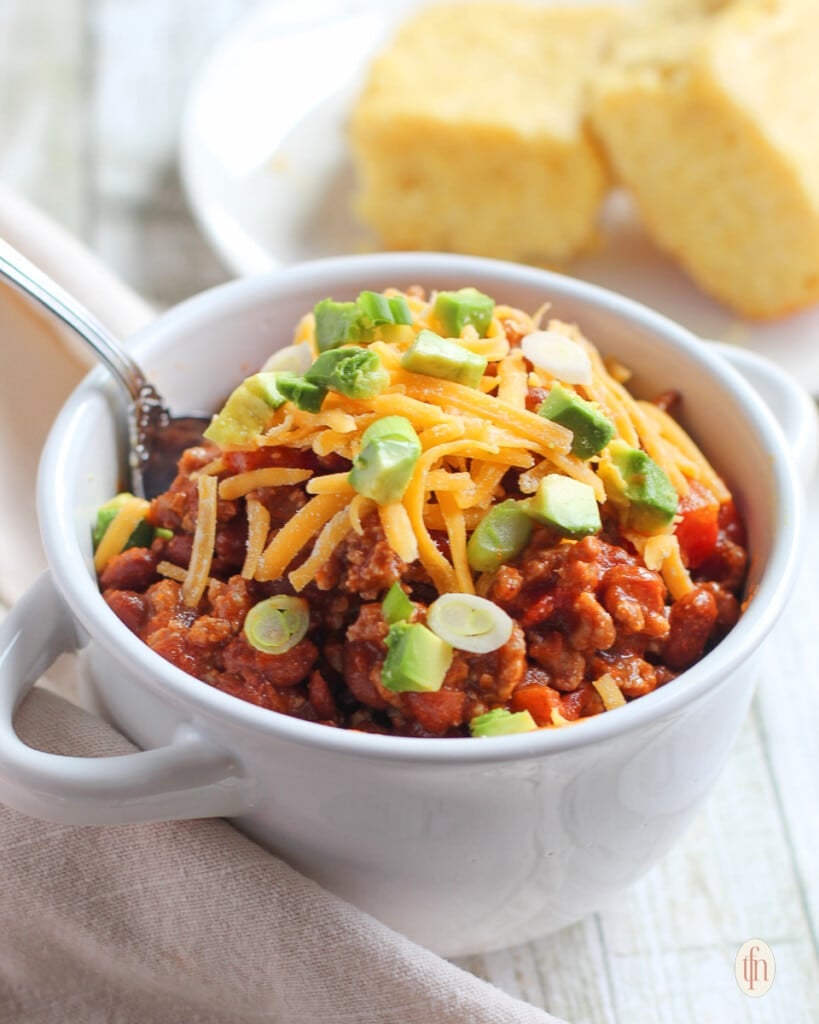 Chili con carne and a spoon in a small bowl with 2 pieces of cornbread in the background.
