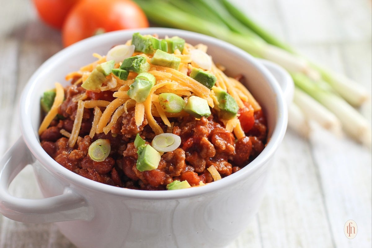 Serving of Dutch oven chili in a white bowl, garnished with grated cheddar cheese and chopped green onions.