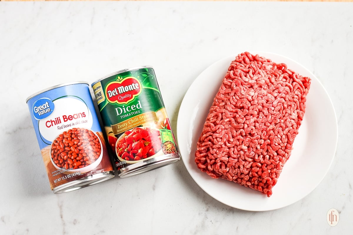 Ingredients for an easy chili recipe on stove; ground beef, canned chili beans, and canned diced tomatoes.