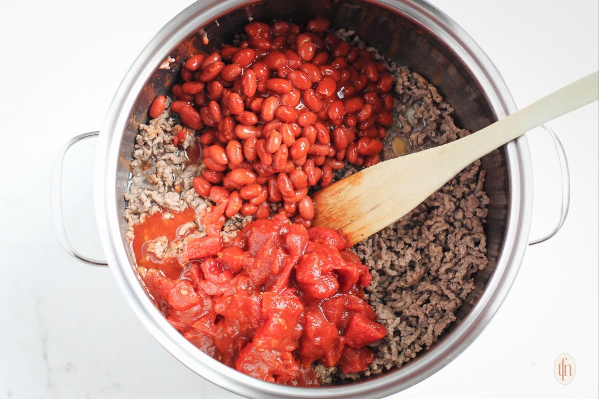 Diced tomatoes, kidney beans, and browned ground beef in a large pot with a wooden spoon.