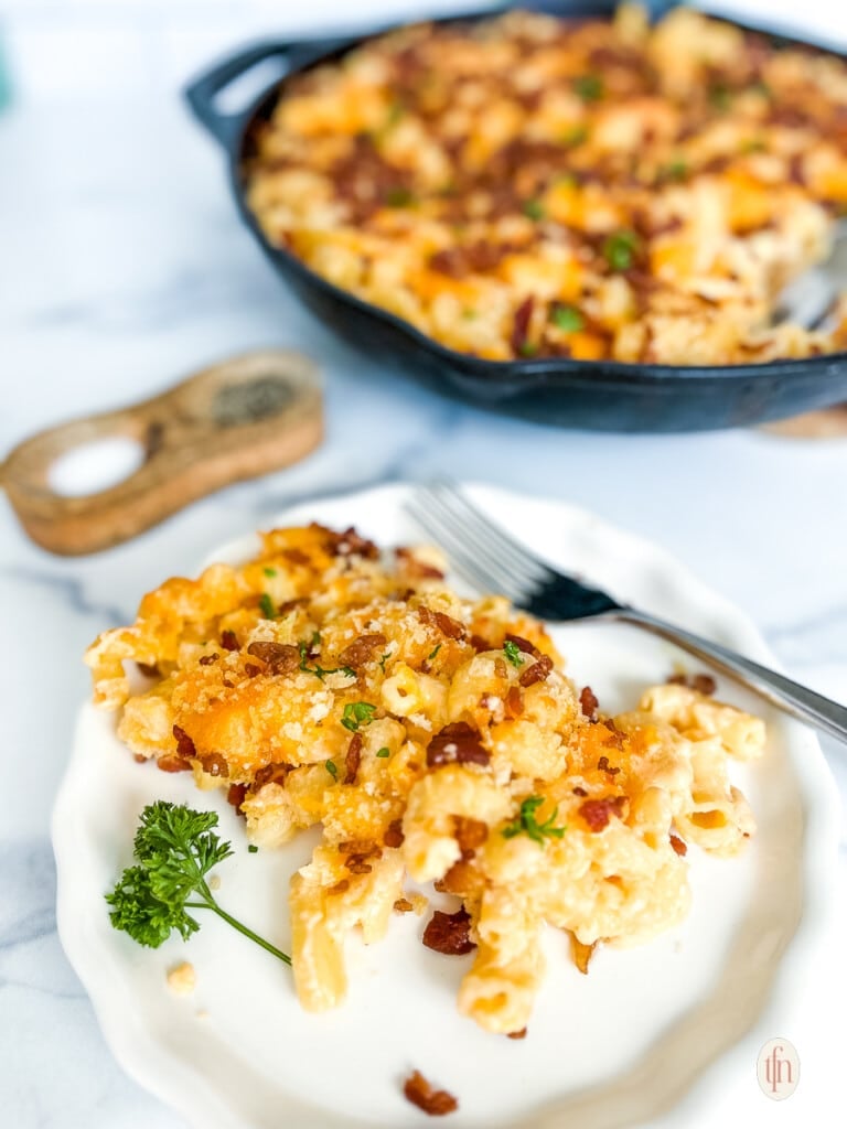 Single serving of pasta with cheese and bacon. Skillet with more of the pasta casserole in the background.