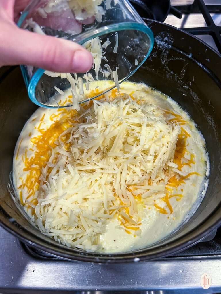 Adding shredded cheddar, gruyere, and white American cheeses to bechamel sauce in a large pot.