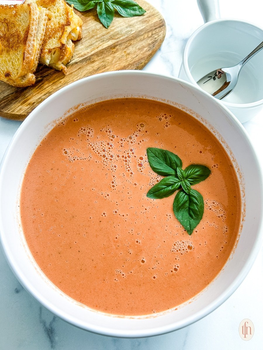 A white bowl filled with tomato soup and a sprig of basil on top.